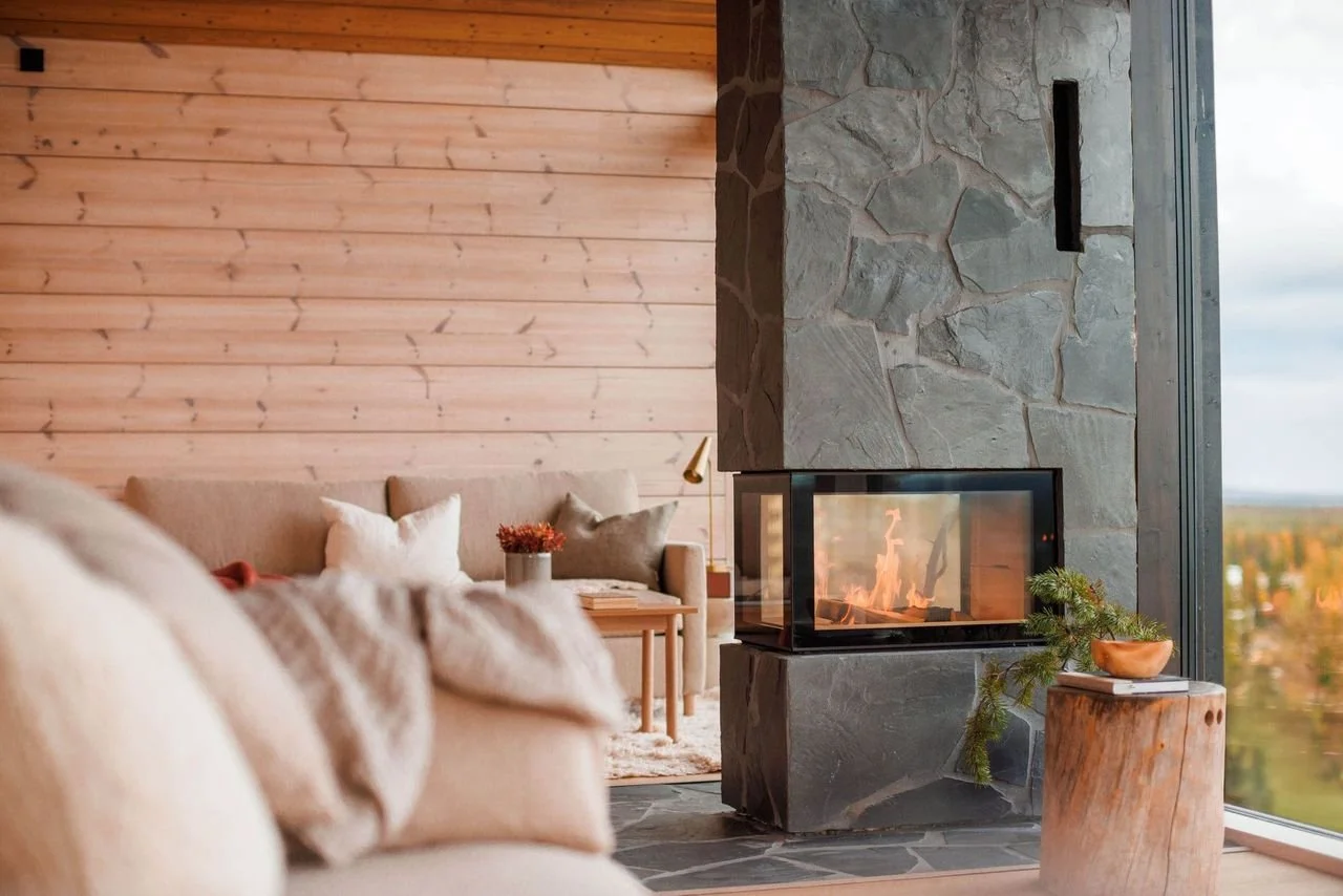 Living room with a beige sofa, pillows, a side table with flowers, a stone fireplace with a glass enclosure, and a large window showing a view of trees and sky outside.