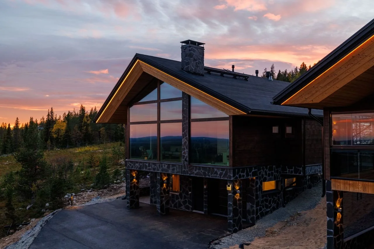 Kide Lodge house with large glass windows and stone exterior, situated in a wooded area during sunset.