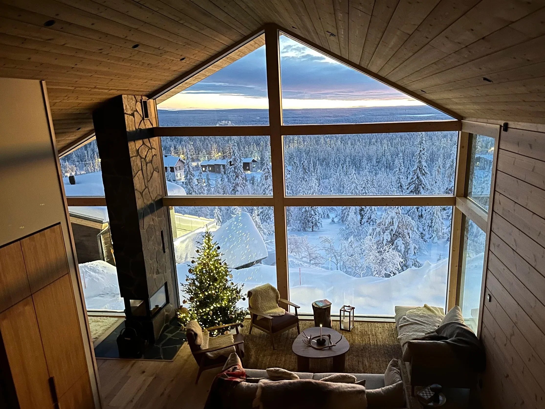 A cozy living room with large floor-to-ceiling windows showcasing a snowy landscape, mountains, and a lake at sunset. The room features a Christmas tree, a fireplace, and comfortable seating.