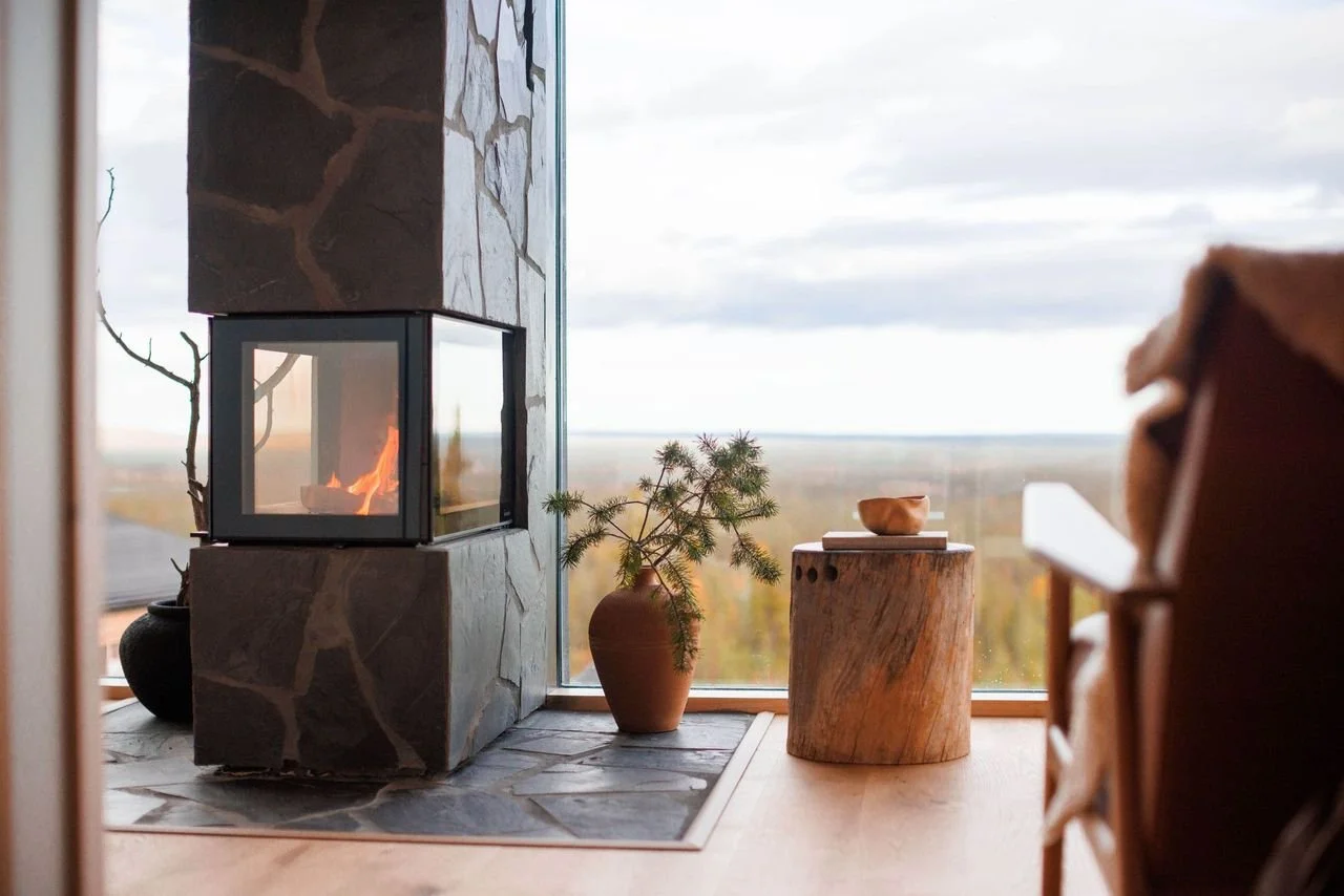 Living room corner with a fireplace, potted plant, and wooden side table near a large window showing an outdoor landscape.