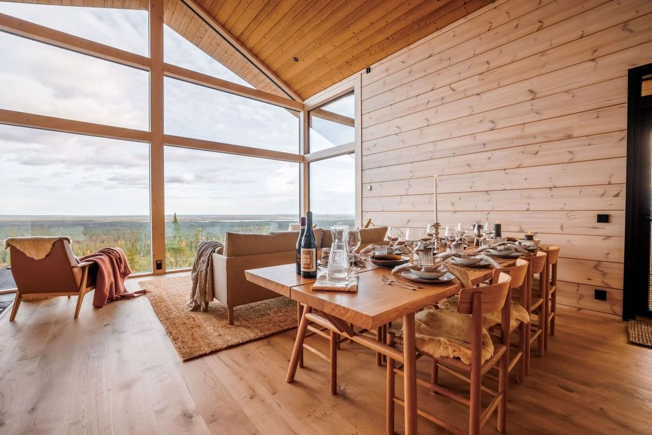 A dining area  with large windows showcasing a scenic view of trees and sky. The table is set with wine bottles, glassware, plates, and a candle. There are chairs with fur throws and a cozy armchair with an orange throw blanket. The room has wooden w