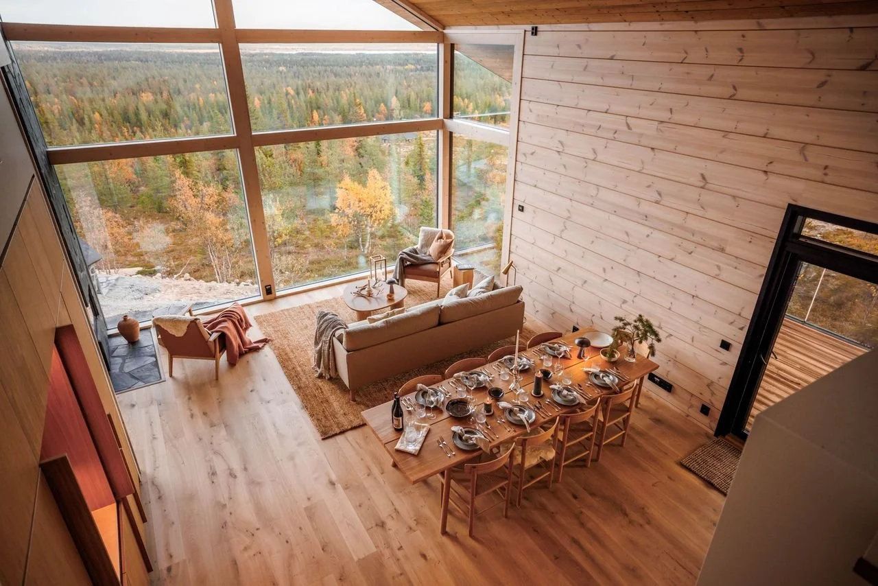 Living and dining area of a modern home with large floor-to-ceiling windows overlooking a forest landscape, wooden walls and flooring, a dining table set for a meal, and a cozy seating area with chairs and a sofa.