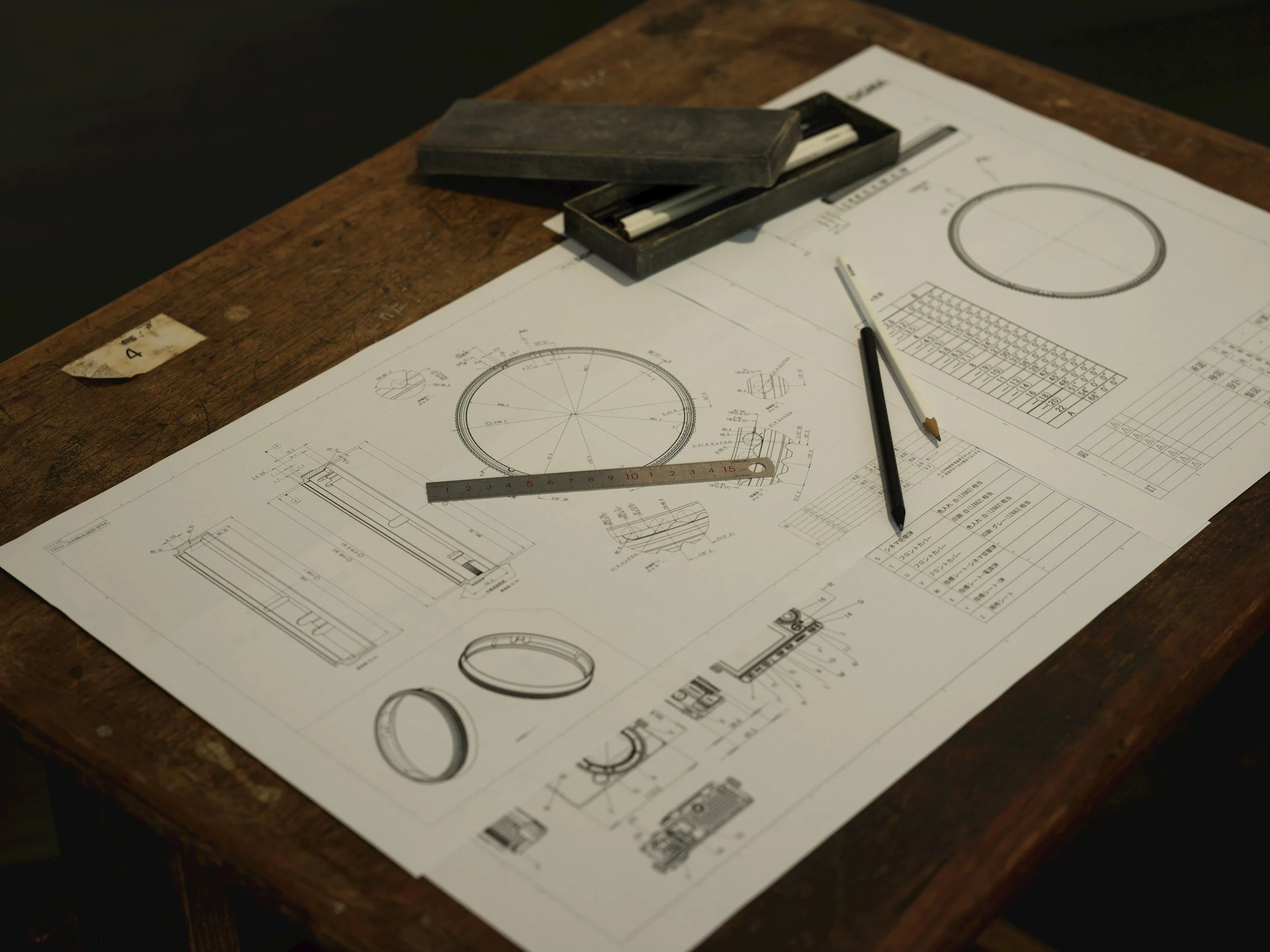 Technical engineering blueprints and drawing tools on a wooden desk, including a ruler, pens, and technical drawings of circular and rectangular mechanical parts.