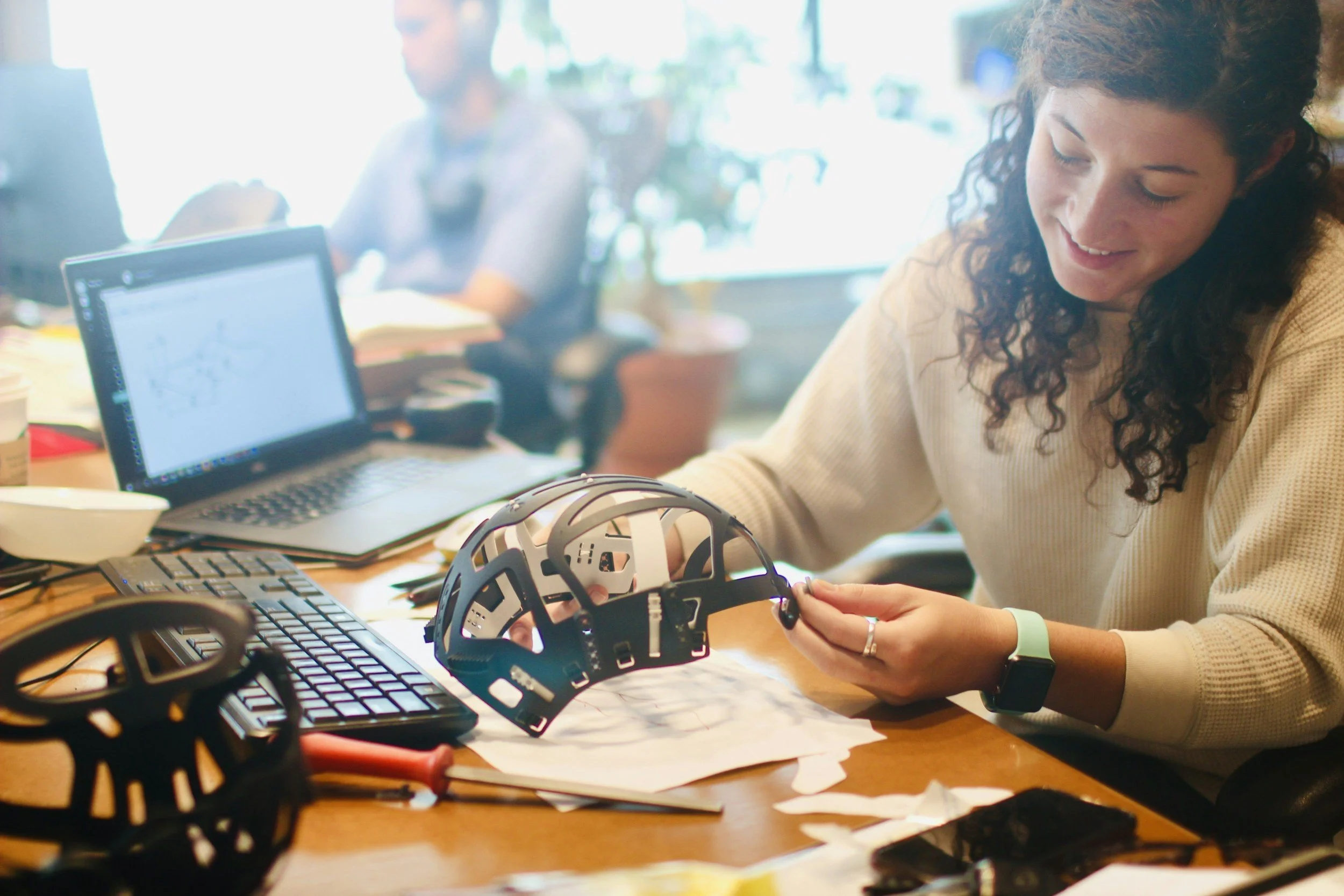 A woman with curly hair wearing a beige sweater and a smartwatch is working on assembling a black robotic helmet at a cluttered desk with a laptop, keyboard, papers, and tools, while smiling.