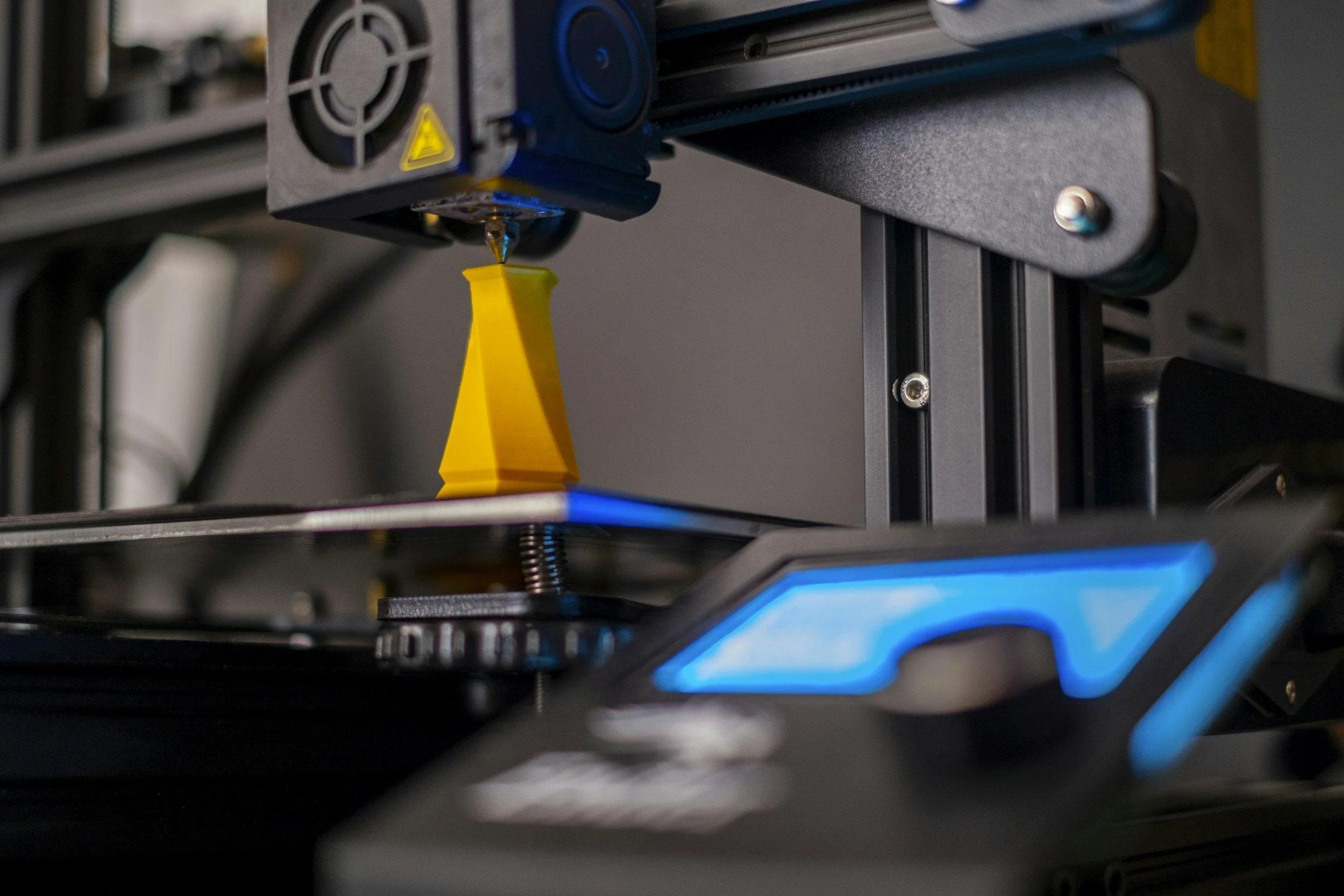 Close-up of a 3D printer extruder nozzle depositing yellow filament onto a print bed.