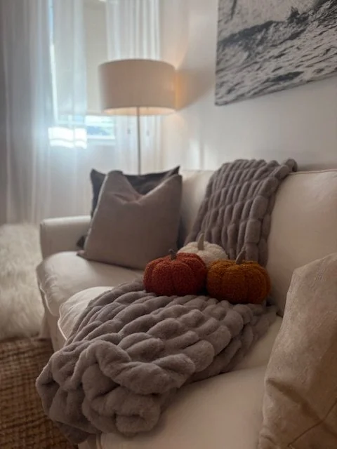 Cozy living room with a beige couch, decorative pillows, a knitted blanket, pumpkins, and a floor lamp near a window with white curtains.