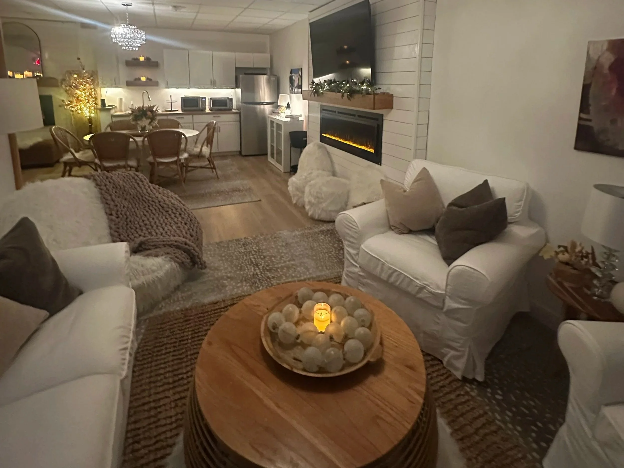 Cozy living room with white sofas, a wooden coffee table with decorative spheres and candle, and a fireplace with holiday decor. In the background, an open kitchen and dining area with four chairs around a table, a chandelier, and a large TV above the fireplace.