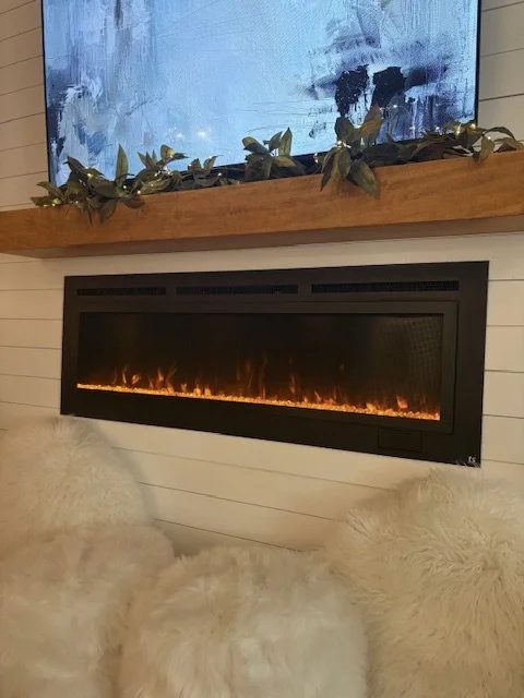 Modern living room fireplace with a black rectangular electric insert, surrounded by cozy fur-covered stools or cushions, and a wall-mounted flat-screen TV above, with a wooden mantle decorated with green leaves.
