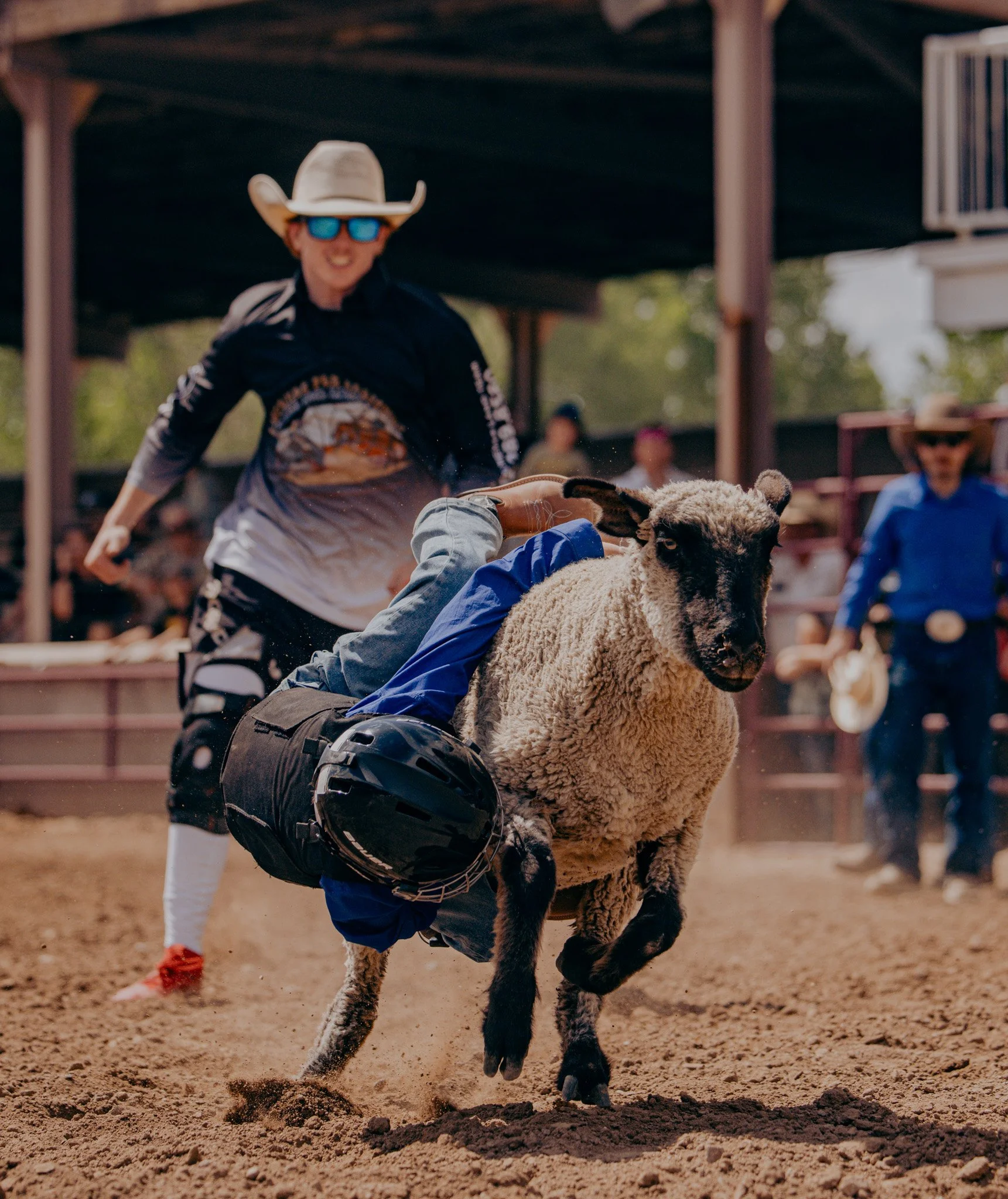 A person wearing a cowboy hat and sunglasses riding a sheep in a rodeo arena while another person in the background watches.