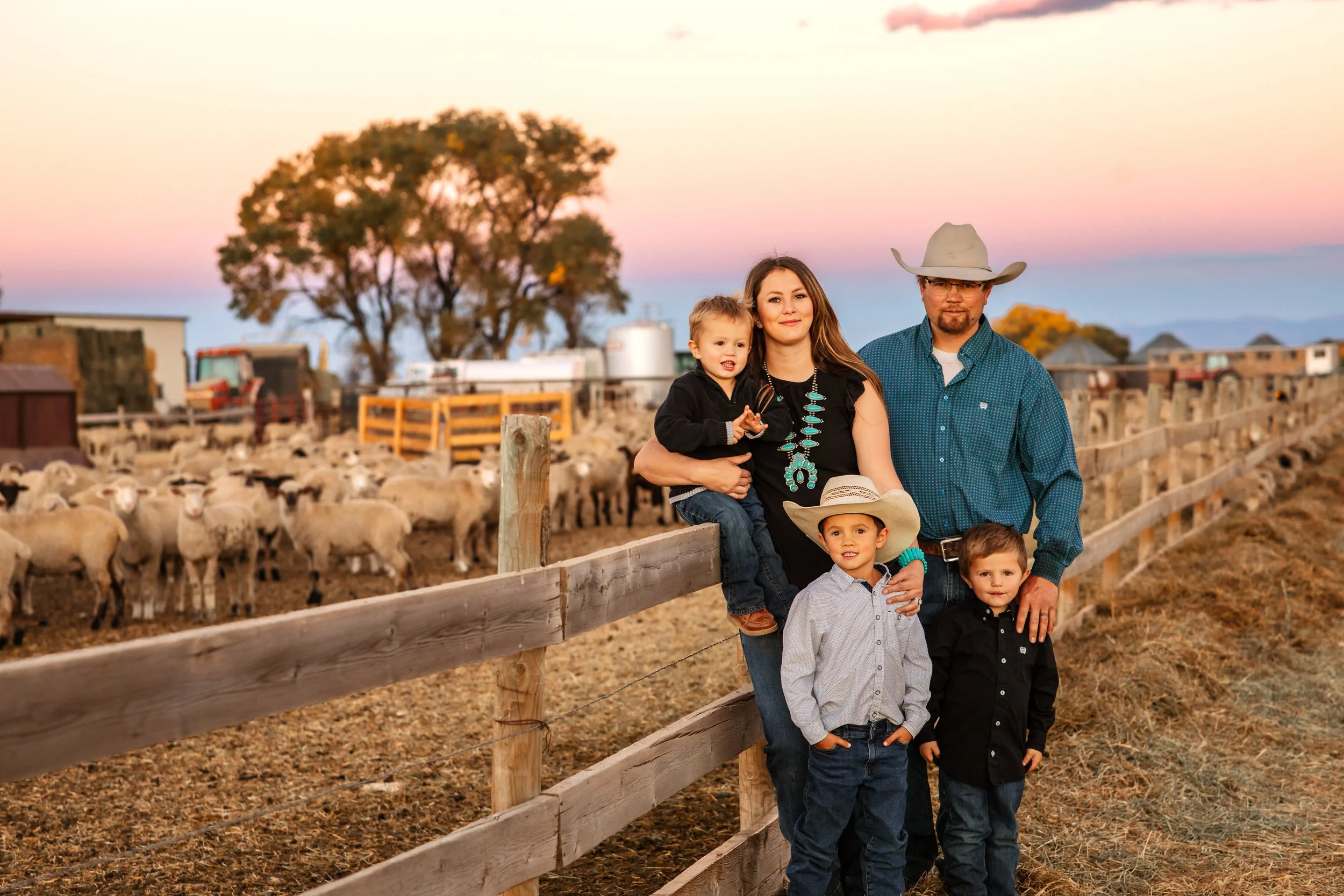 A family of six stands by a wooden fence in a rural farm setting at sunset. The background features a large sheep pasture, farm buildings, and a large tree. The family includes a man wearing a cowboy hat and glasses, a woman with long hair wearing a black shirt, and four young children dressed casually. The sky is colorful with pink and purple hues.