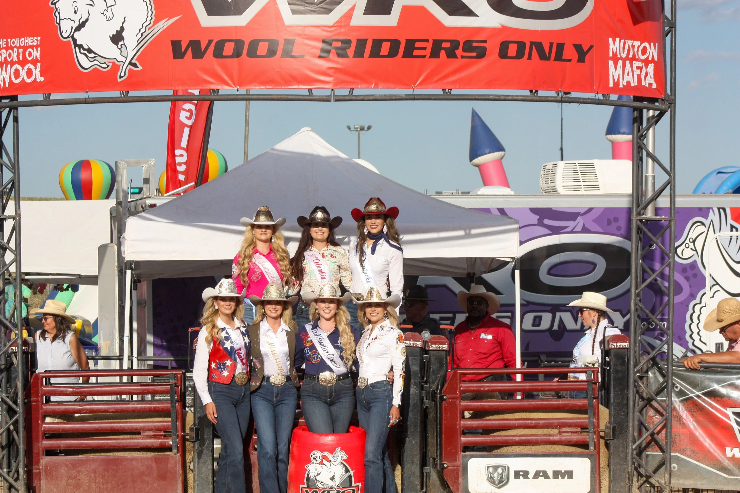Group of women dressed in western and rodeo attire, standing on a rodeo parade float with a red banner overhead that reads 'WoL RIdERS ONLY', with parachute-shaped inflatables and wind balloons in the background.