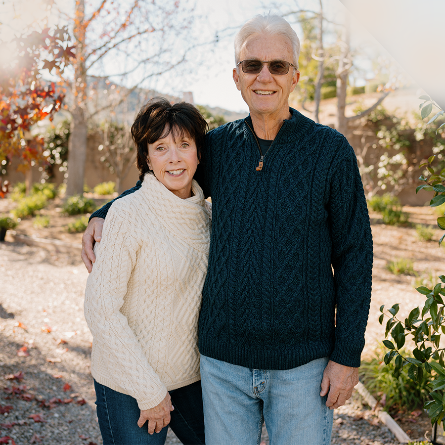 An elderly man and woman standing outdoors on a sunny day, smiling. The man has white hair, sunglasses, and a dark blue sweater. The woman has short brown hair and is wearing a cream-colored sweater. They are hugging and standing in a garden with trees and plants.
