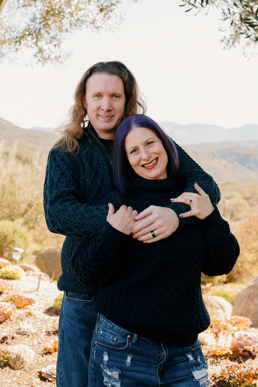 A smiling couple outdoors, with mountains and trees in the background. The man has long, wavy hair and is wearing a dark sweater, while the woman has purple hair and is wearing a black sweater and ripped jeans.