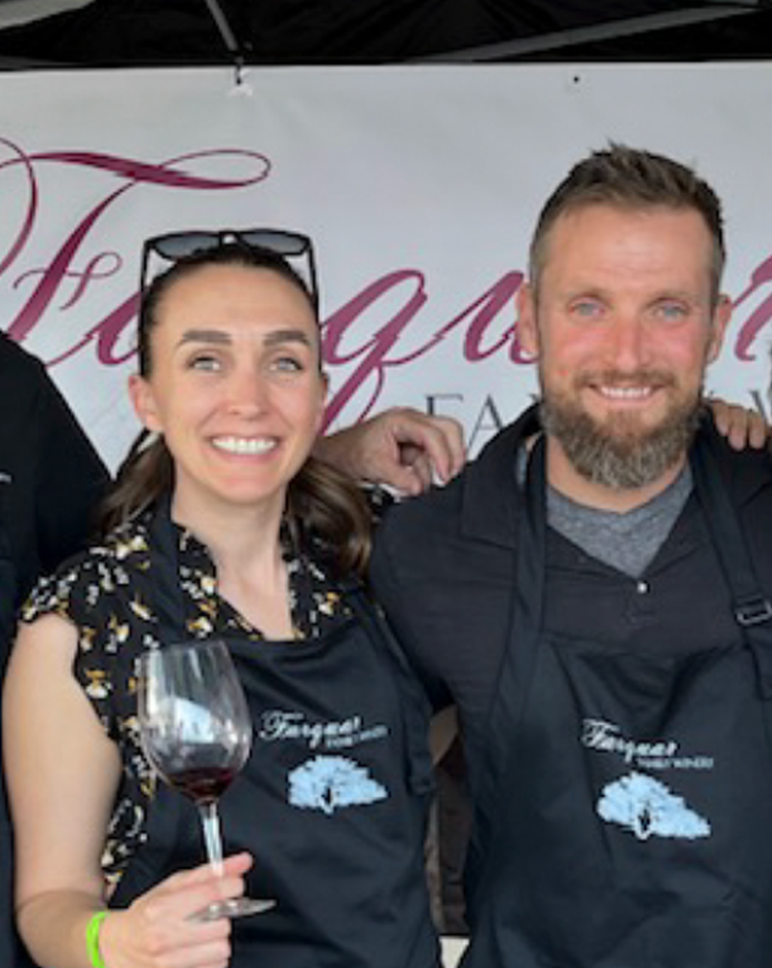 A woman and man smiling at a food event, wearing aprons with a tree logo, woman holding a glass of red wine, background has pink cursive text.
