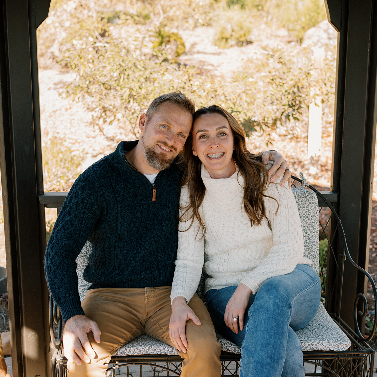 A smiling couple sitting on a bench in a black wooden gazebo outdoors with trees and autumn foliage in the background.