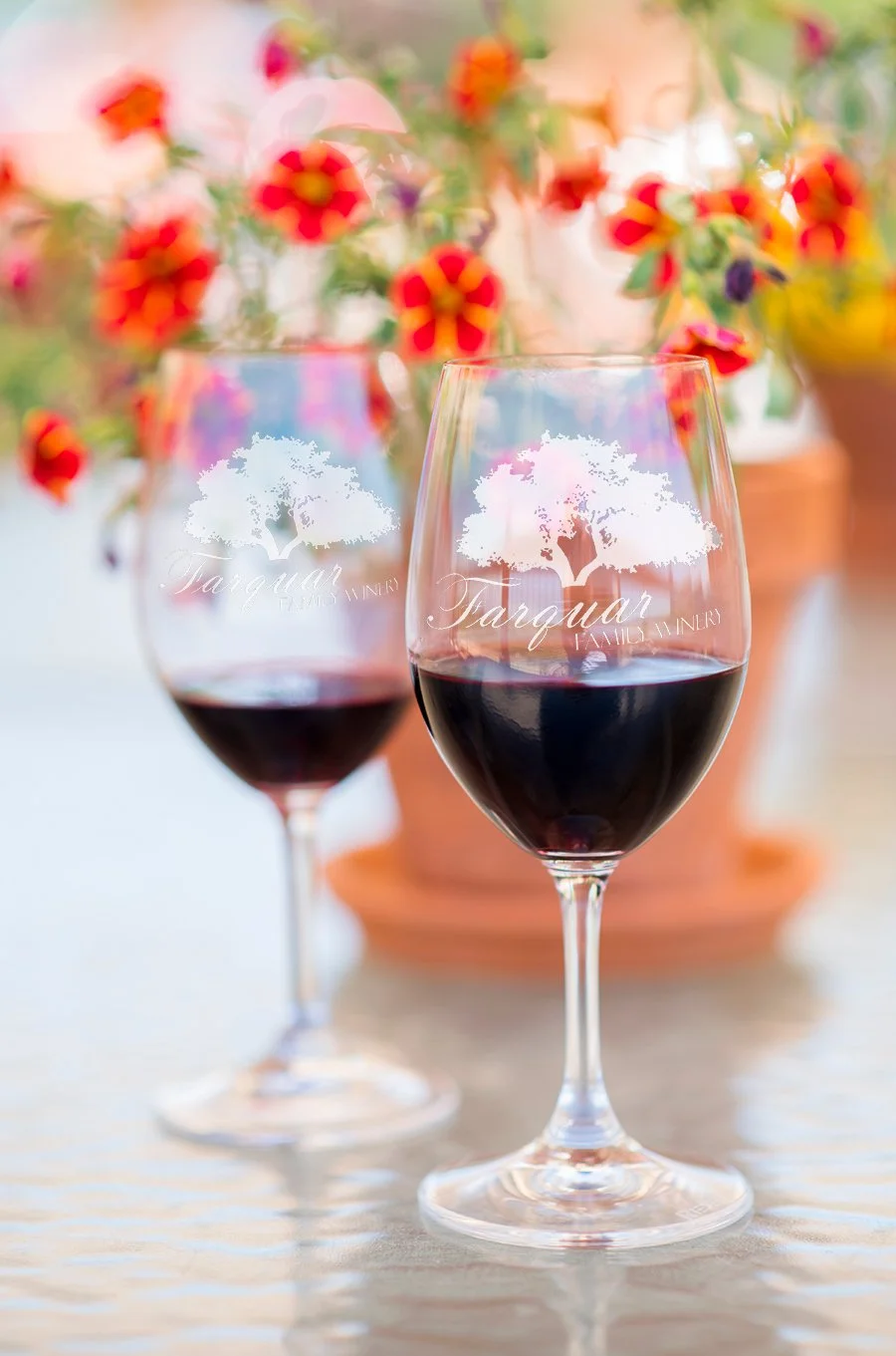 Two wine glasses filled with red wine on a table, with a blurred background of colorful flowers and a terracotta pot.