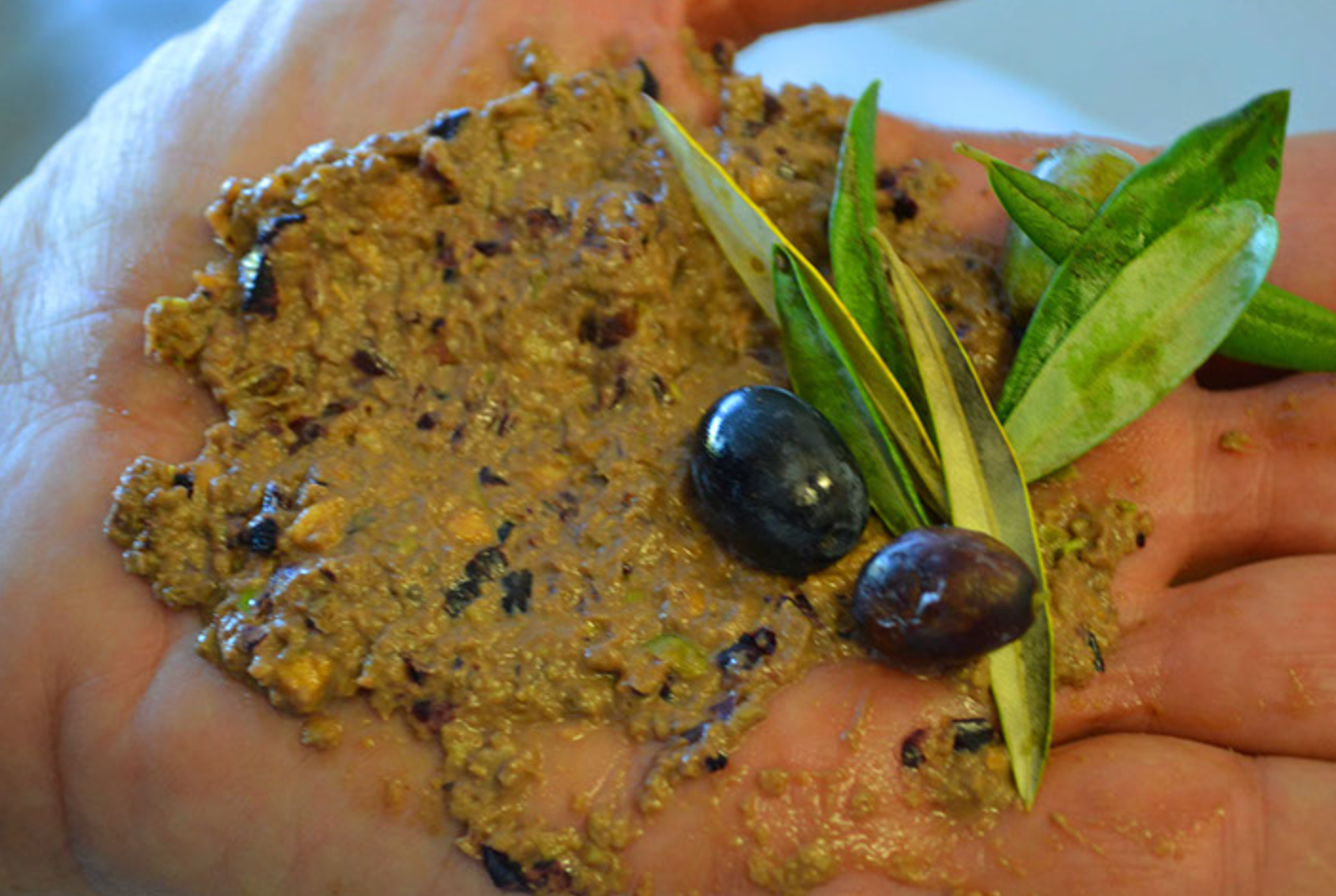 Hand holding a piece of chopped, cooked plantain topped with ground meat, along with a few green leaves and two dark olives.