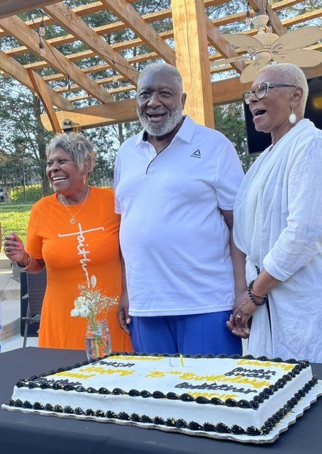 Three older adults smiling and standing behind a large birthday cake outdoors, with a wooden pergola overhead.