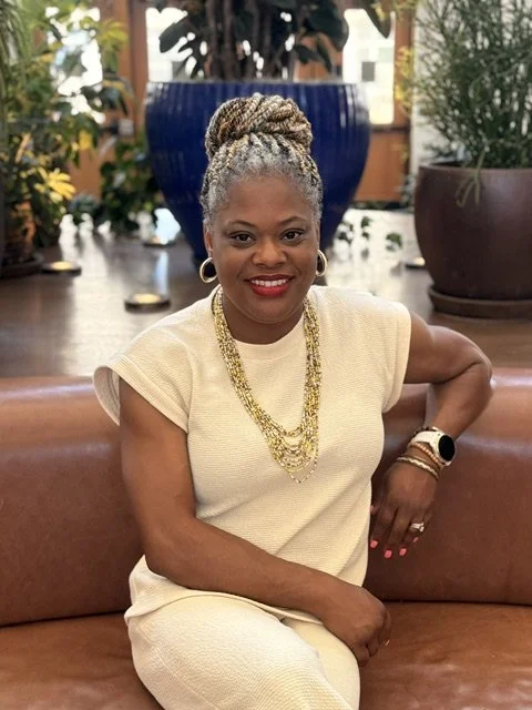 A woman with gray braided hair styled in an updo, wearing a beige dress, gold jewelry, and a smartwatch, sitting on a leather couch in a well-lit indoor space decorated with plants and large pots.