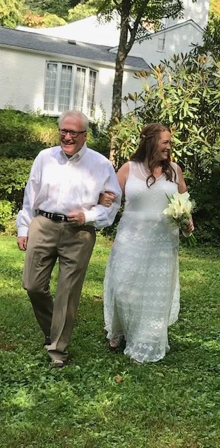 A bride in a white wedding dress holding a bouquet, walking arm-in-arm with an older man dressed in a white shirt and khaki pants, outdoors in a green yard with trees and a white house in the background.
