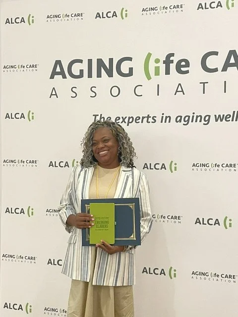 A smiling woman holding a certificate or award in front of a backdrop with the logos of ALCA: Aging Life Care Association and the phrase 'The experts in aging well'.