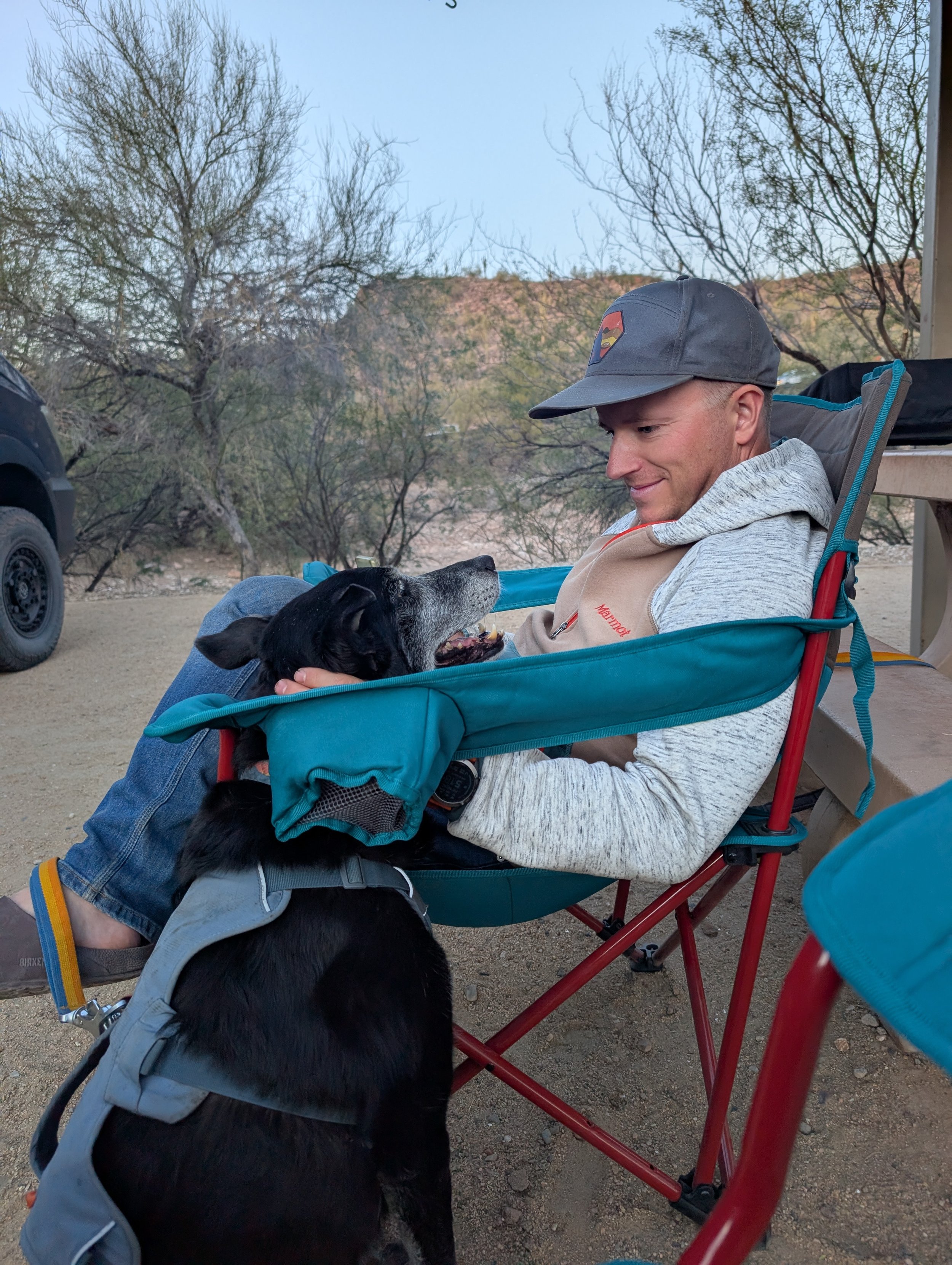 A man sitting in a camping chair, smiling and holding a black and white puppy in his lap with a backpack nearby. Outdoors with leafless trees and mountains in the background.