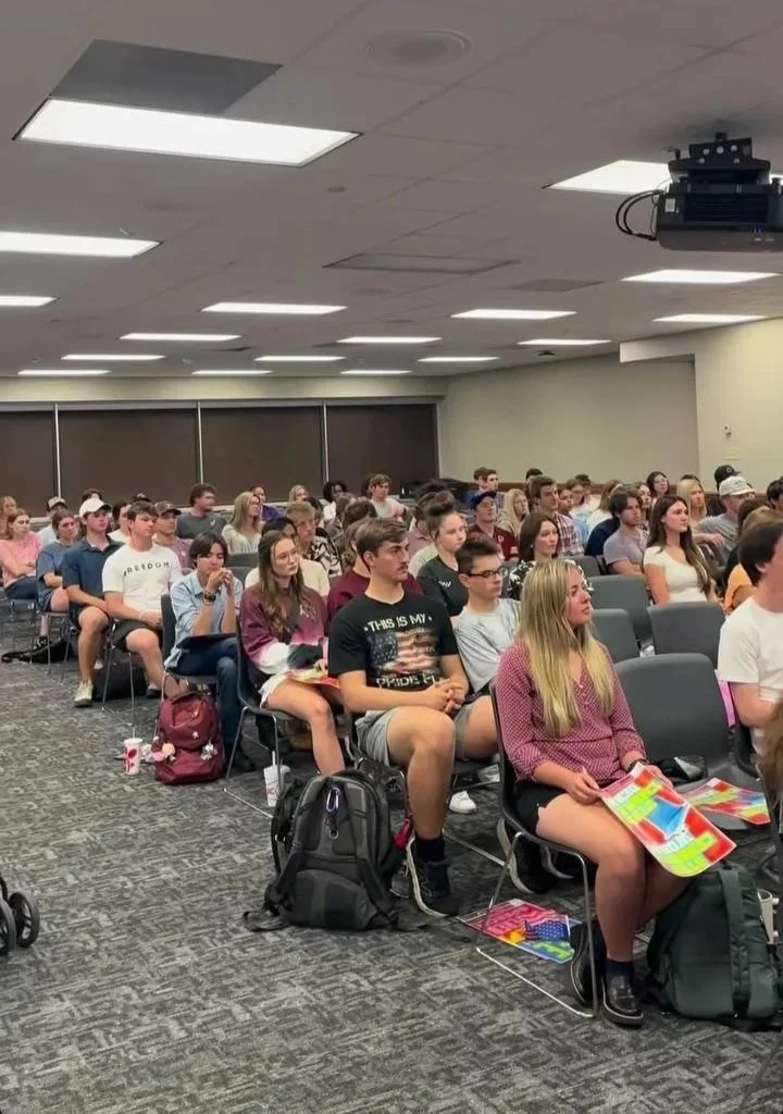 A large group of students sits in a classroom, attentively listening to a lecture or presentation. They are seated in rows with backpacks and notebooks, under bright ceiling lights in a modern classroom or conference room.