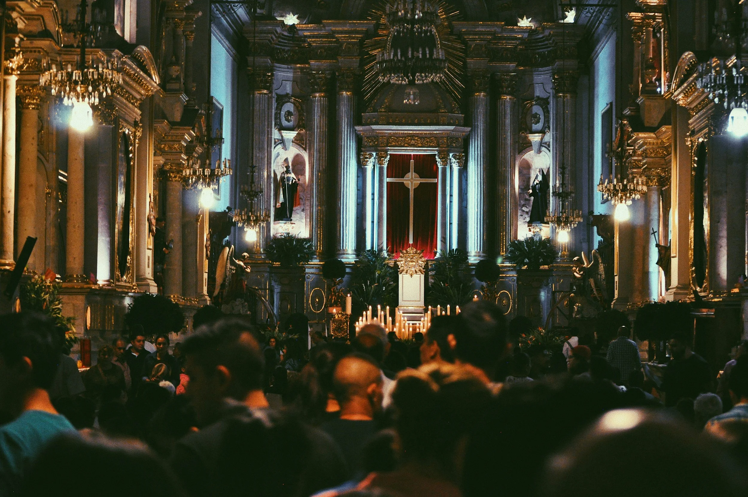 Interior of a church filled with people, featuring ornate gold detailing, statues, candles, and a large cross at the altar.