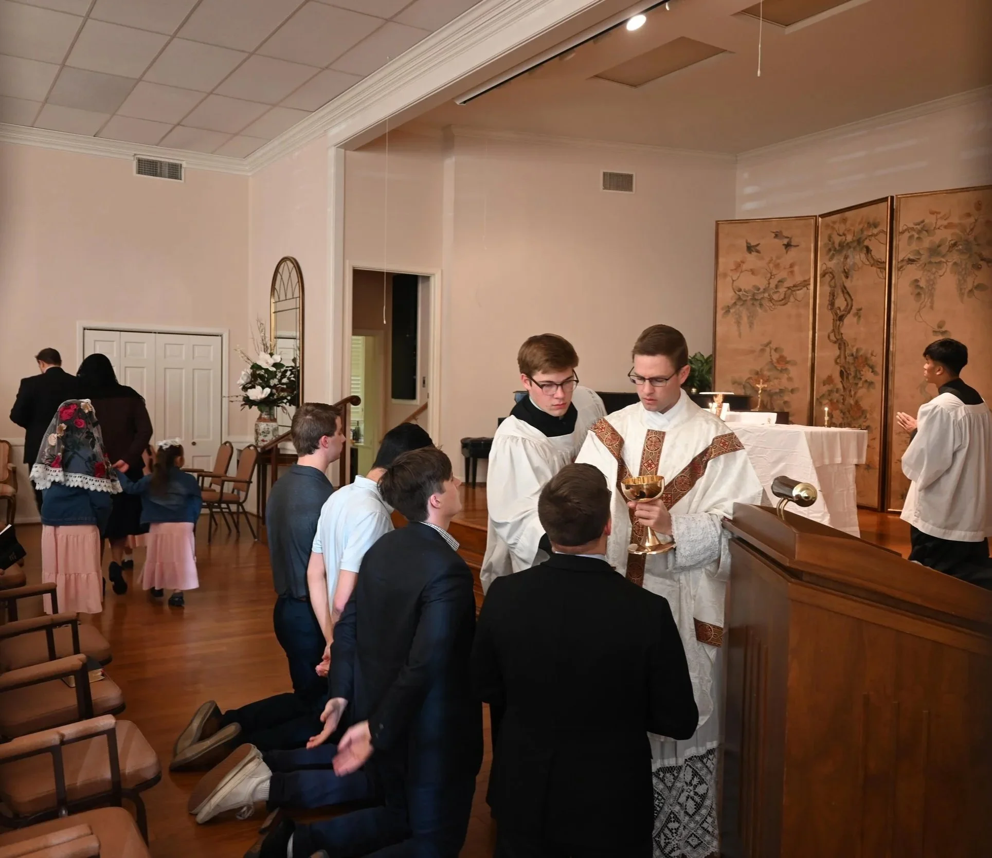 A religious ceremony taking place inside a church, with a priest and altar servers conducting a ritual before kneeling individuals, and a group of children and adults in attendance.
