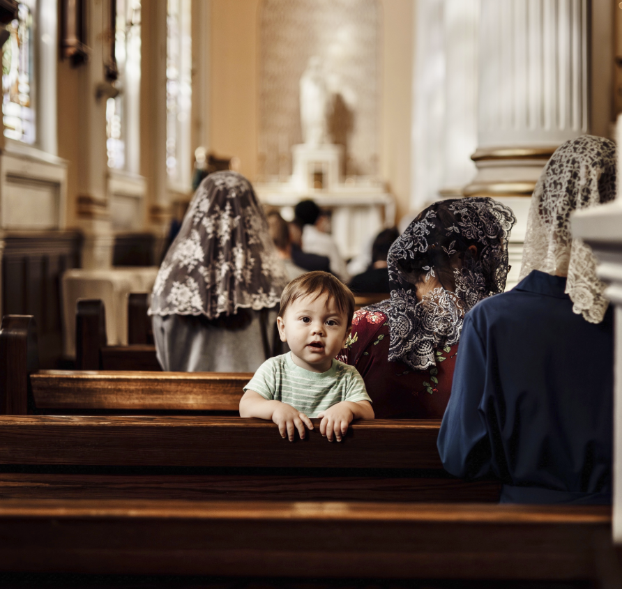 A young child looks towards the camera while sitting in a church pew, surrounded by women wearing lace head coverings.