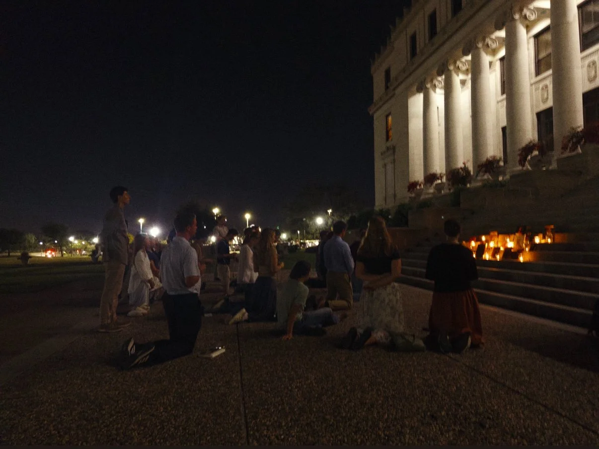 People kneeling and sitting on the steps of a building at night, participating in a candlelight vigil.
