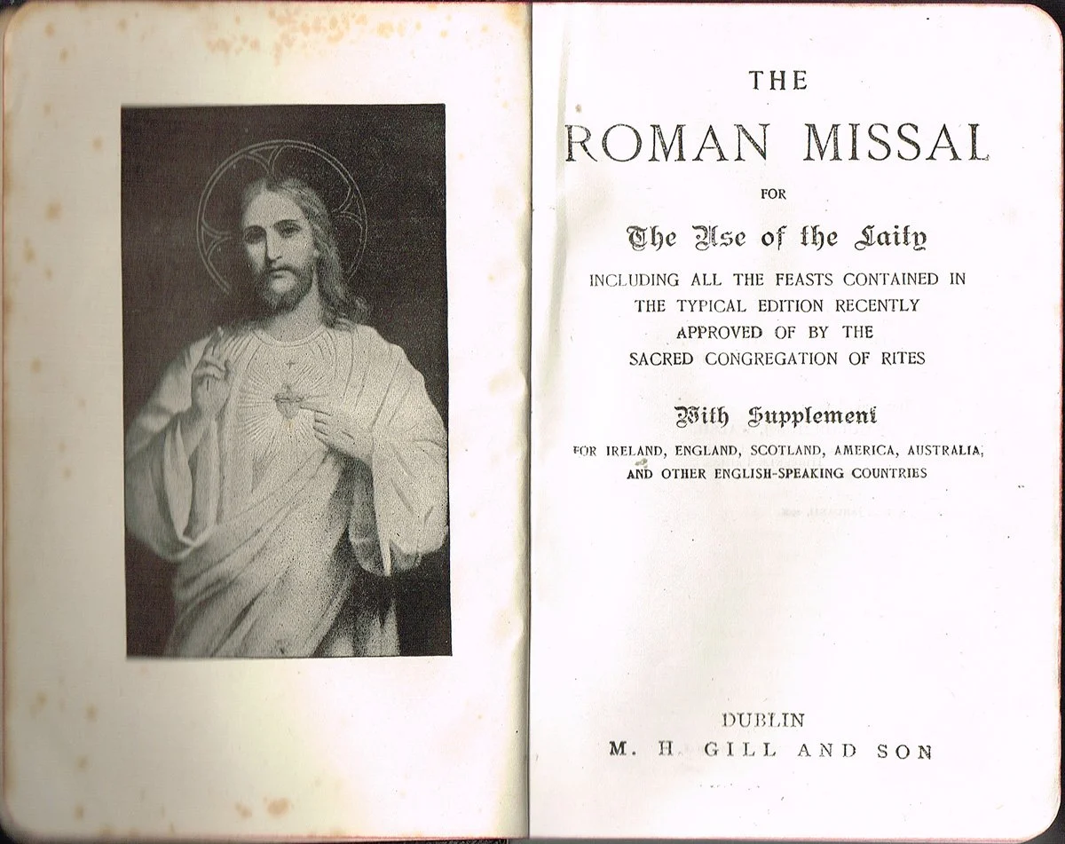 A black and white illustration of Jesus Christ with a halo, holding his right hand up in blessing and pointing to his chest with his left hand. Next to the illustration is a title page of a book titled 'The Roman Missal for the Use of the Laity' with publication details from Dublin, M. H. Gill and Son.