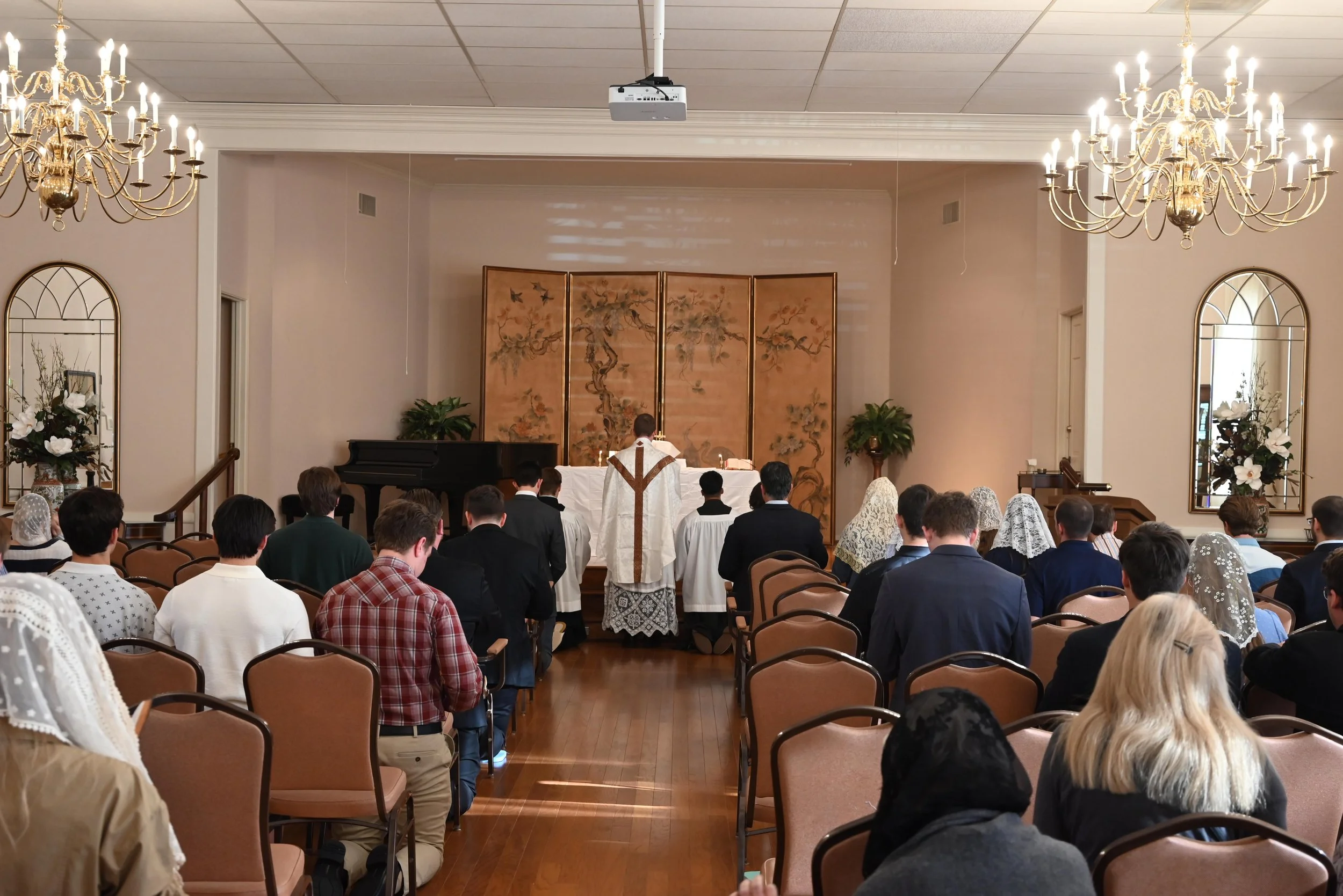 A church congregation attending a religious service with a priest at the altar, with chandeliers, mirrors, and floral arrangements in the background.