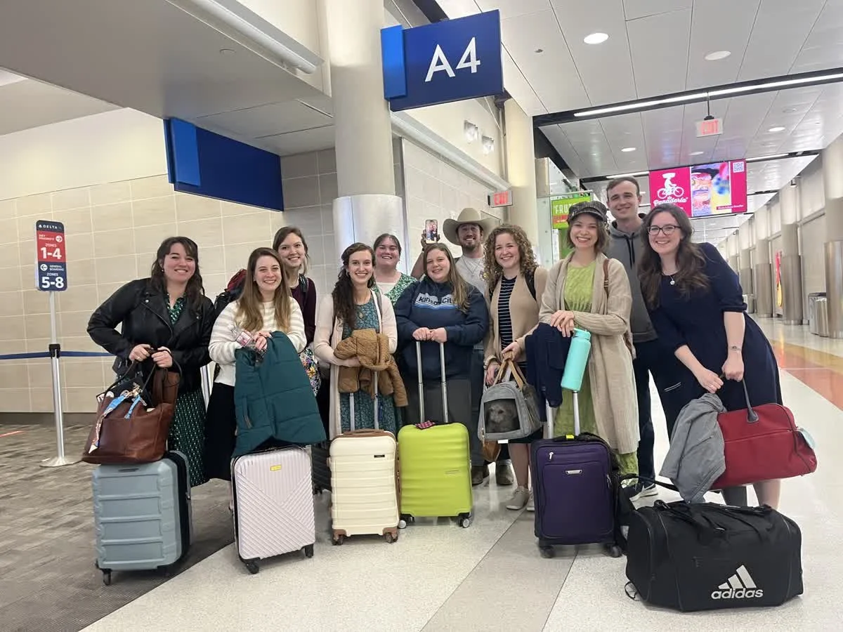 Group of people at an airport terminal, standing in front of gate A4, with luggage and backpacks, smiling and posing for a photo.