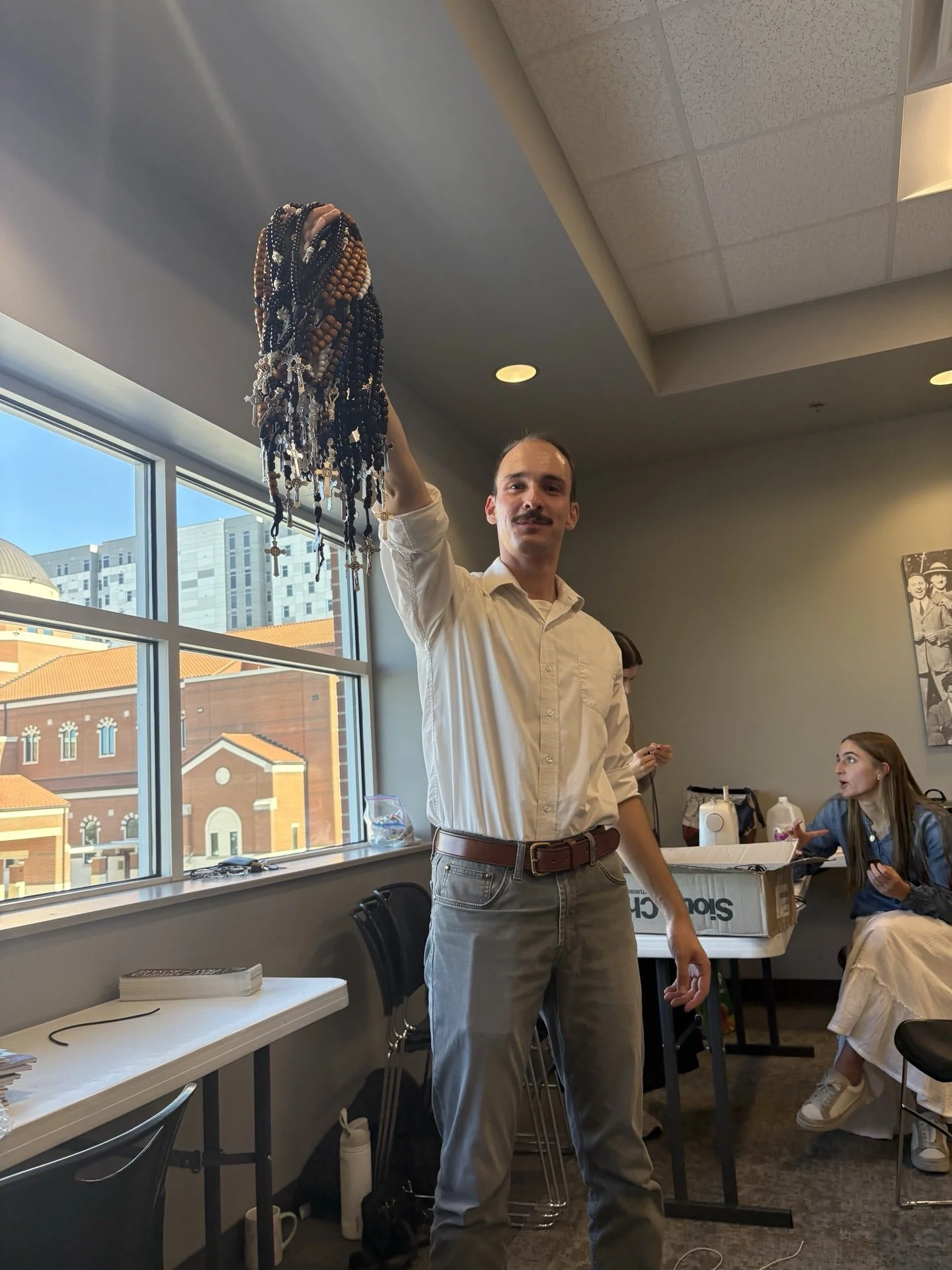 A man with a mustache, wearing a white shirt and gray pants, is holding a large bunch of rosary beads or prayer beads overhead inside a room with large windows and a cityscape view.