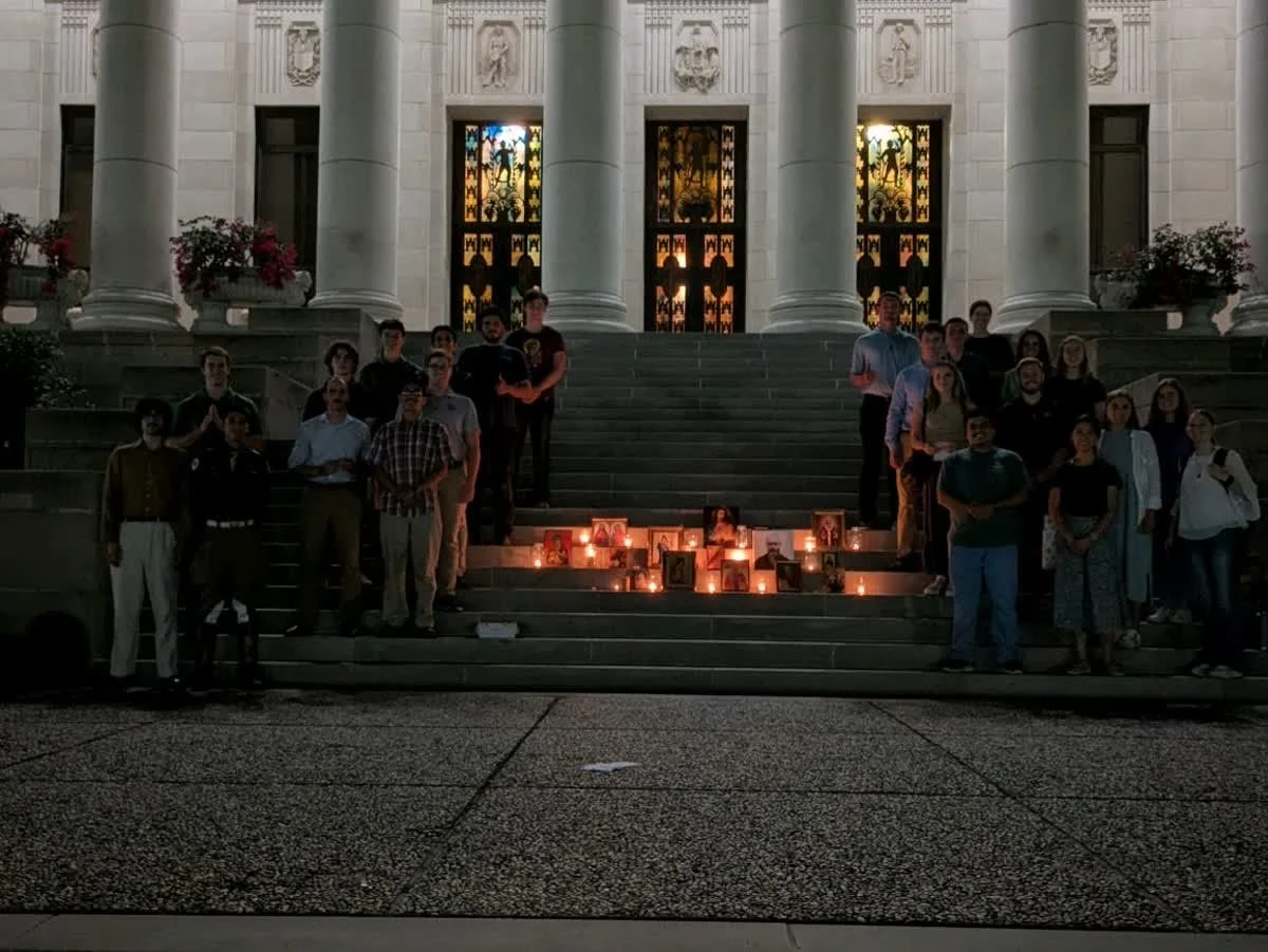 Group of people gathered on steps in front of a building with large columns, holding candles and portraits, at sunset.