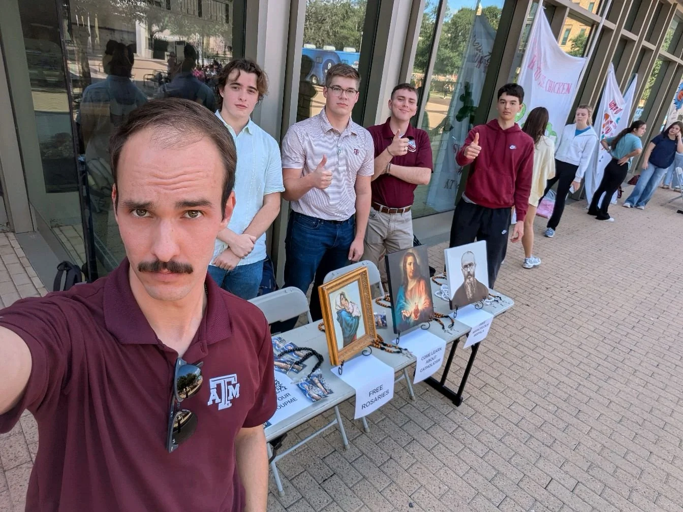 Group of young people standing behind a table with religious images and rosaries outside a building, some giving thumbs up, during a daytime event.