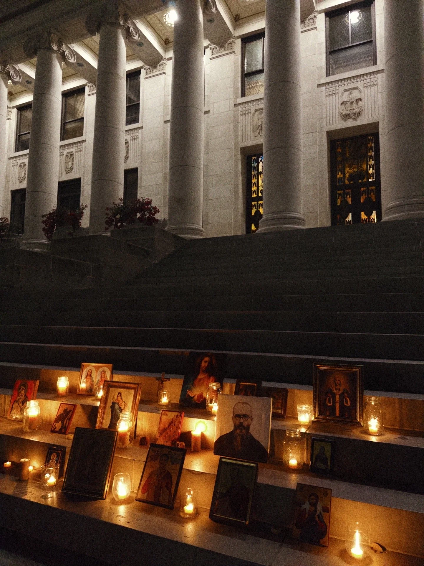 Nighttime scene of a church steps display with candles and religious icons, including portraits of Jesus and saints, illuminated softly in front of a grand building with tall columns and decorative windows.