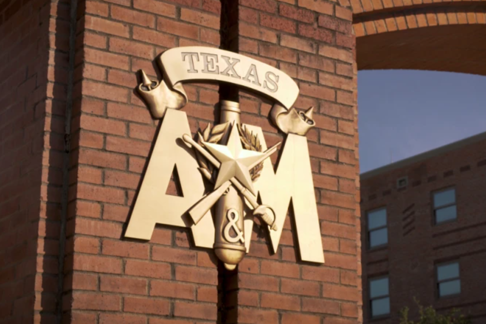 Metal sign on a brick wall showing 'TEXAS A&M' with a star, rifle, and other symbols.