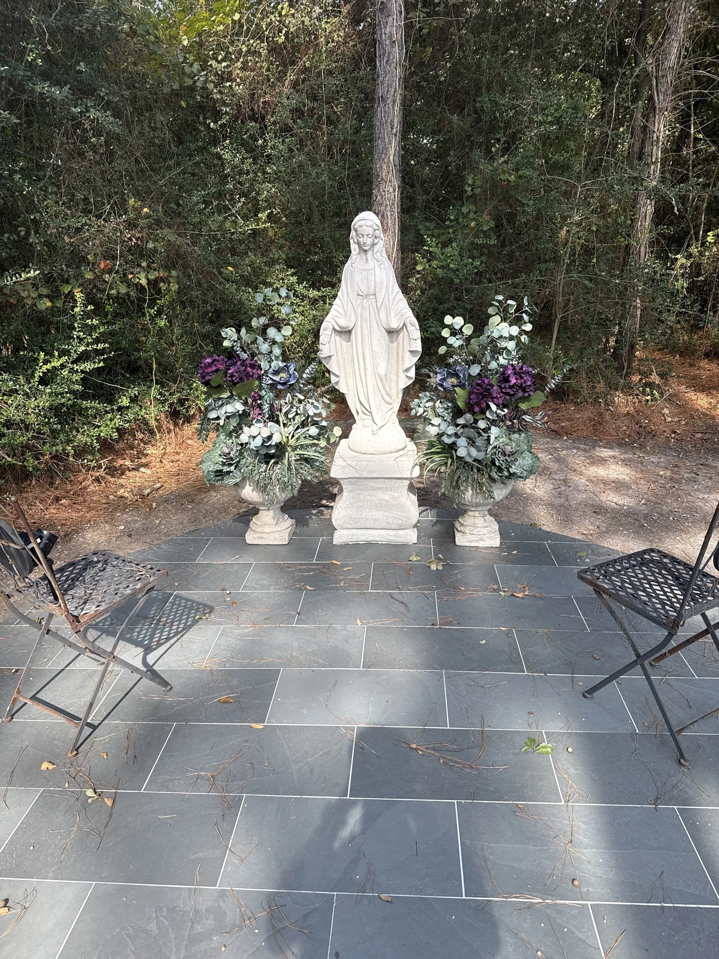 A white religious statue of the Virgin Mary on a tiled outdoor area, flanked by two large flower arrangements with purple and green foliage, surrounded by trees and bushes.
