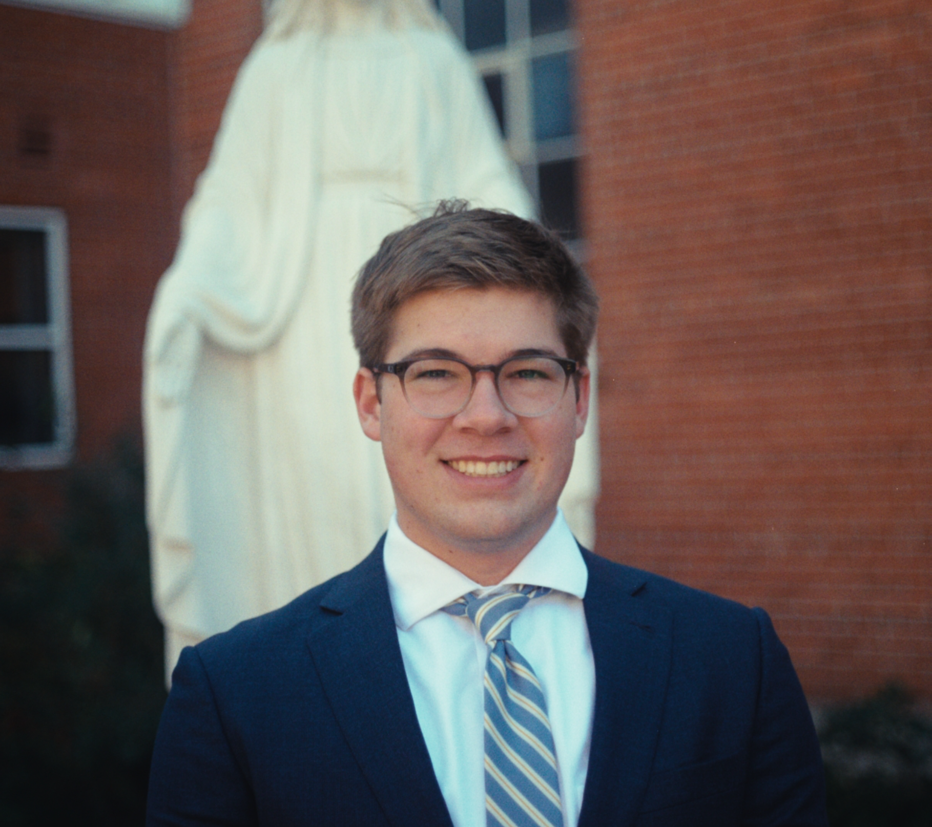 A man with dark curly hair, wearing clear glasses, a nose ring, and a black sweater, sitting in a room with white walls and a green plant in the background.