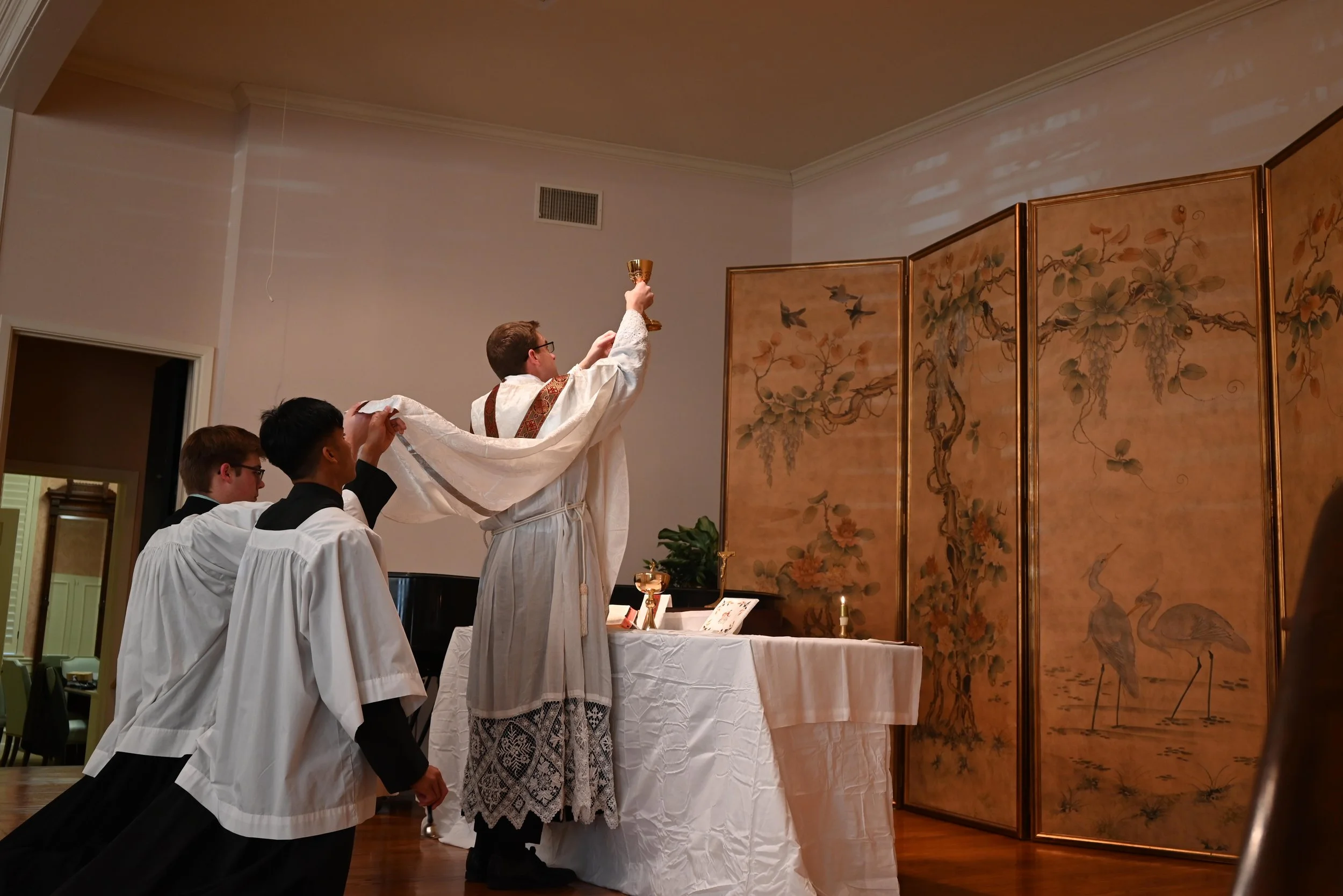 A religious ceremony where the priest or pastor is holding a chalice high, with three altar servers in robes kneeling and supporting him, in front of a decorated folding screen with floral and bird motifs.
