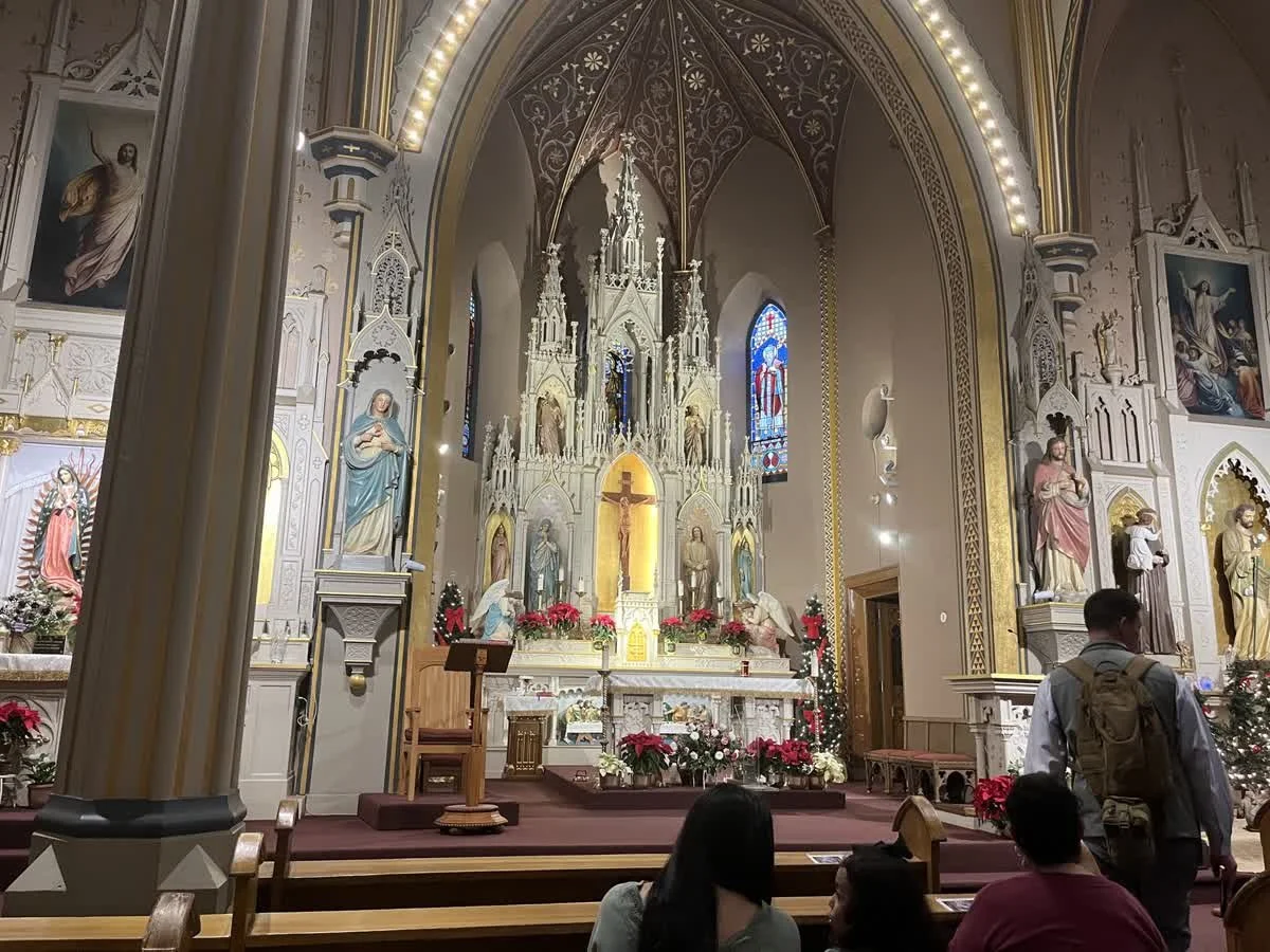 Interior of a church with an ornate altar decorated with poinsettias, statues, and stained glass windows. Several people are seated or standing near the altar.