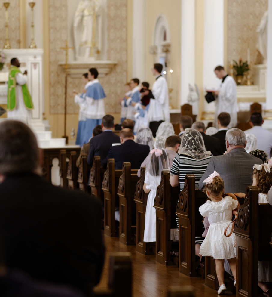 People attending a religious ceremony in a church, with altar and clergy in the background, children dressed in white and adults seated in pews.
