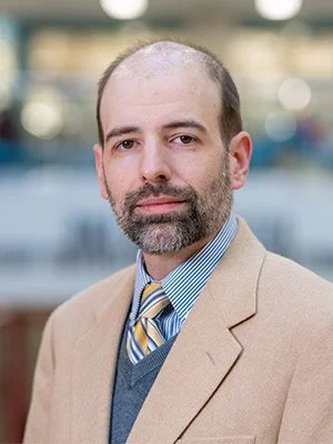 A man with a beard and receding hairline wearing a tan blazer, blue striped shirt, and yellow-striped tie, standing outdoors with a blurred cityscape background.