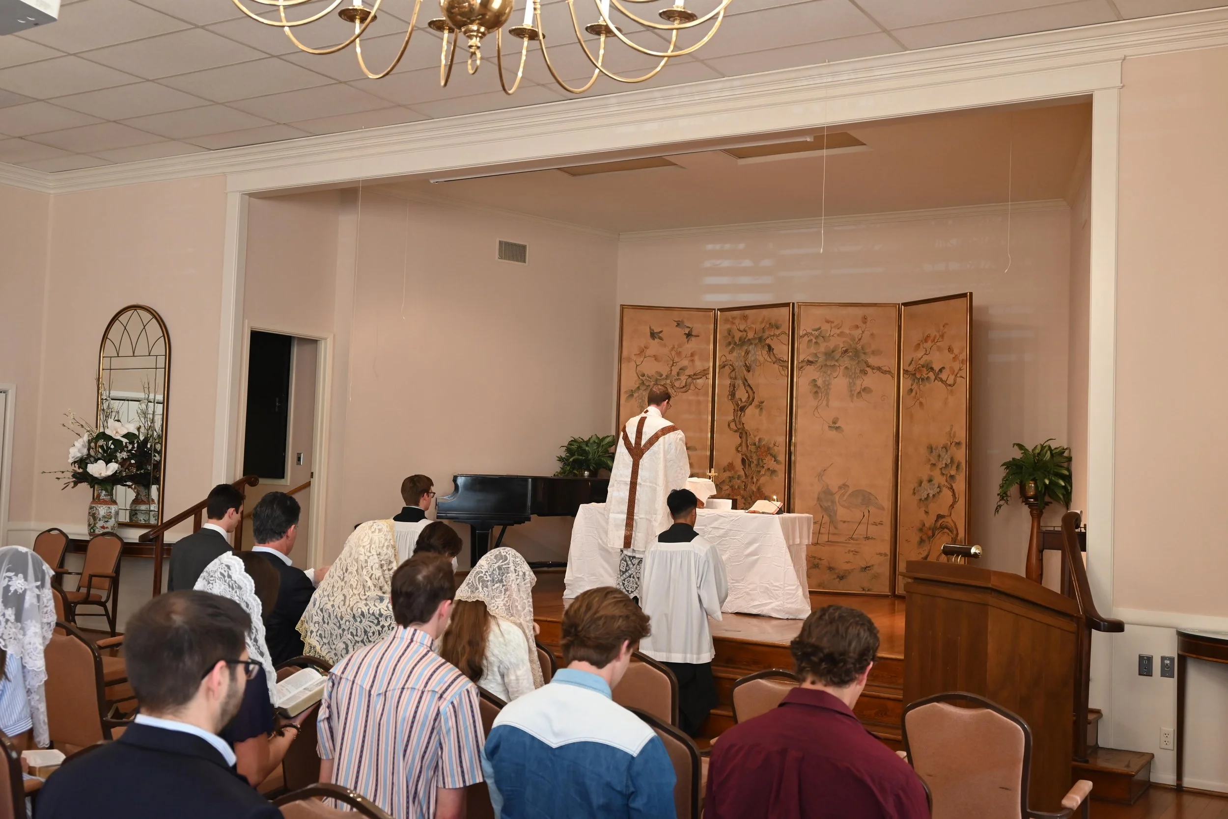 A religious ceremony in a church or chapel with a priest at the altar and congregation seated in pews. The priest is wearing white vestments with a brown stole, facing the altar which has a decorative screen with traditional Asian artwork. The sanctu