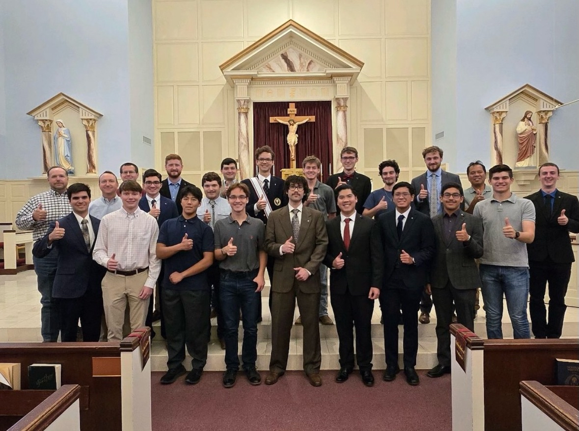 Group of young men and boys standing inside a church, smiling and giving thumbs-up, with a crucifix and religious statues behind them.