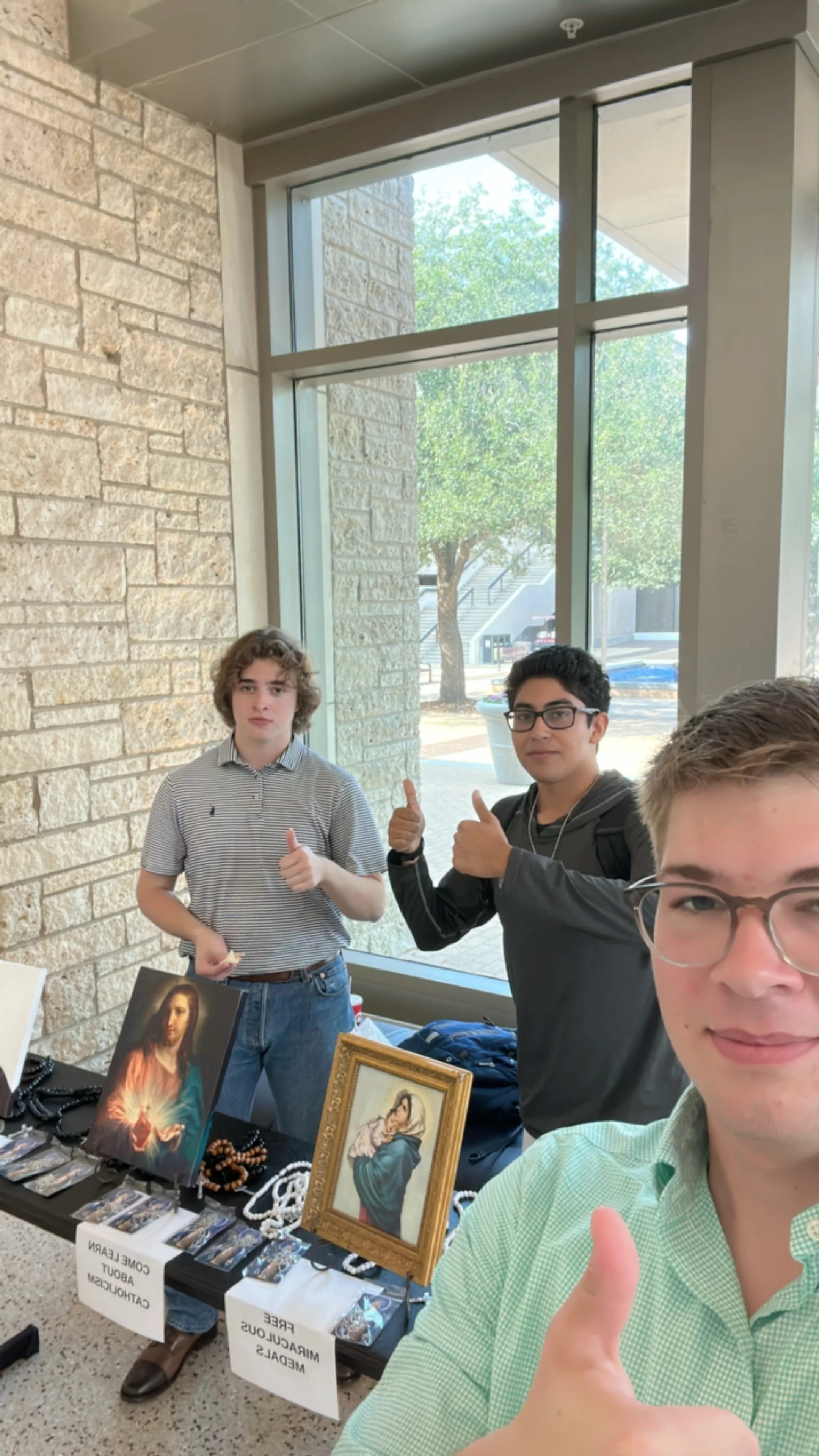 Three young men at a booth displaying religious artwork and jewelry, giving thumbs up, inside a building with large windows.