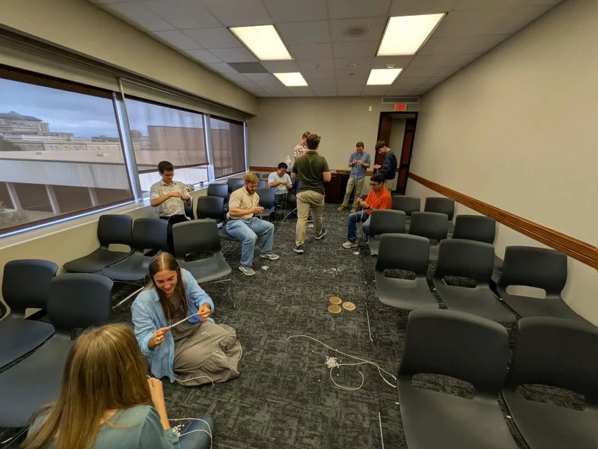 Group of people sitting and standing in a room with chairs, large windows, and a city view, some using electronic devices.