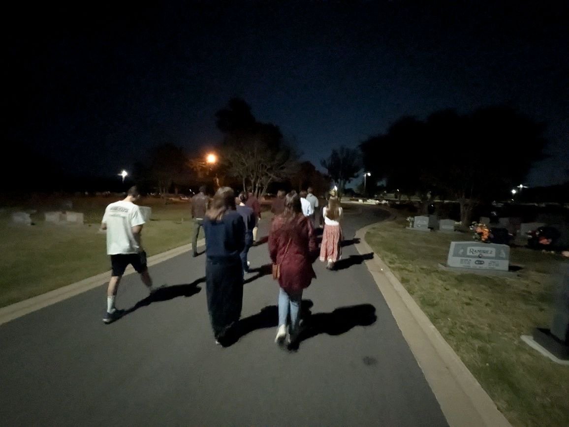 A group of people walking along a paved path in a park at night, illuminated by streetlights, with trees and a gravestone visible on the right side.